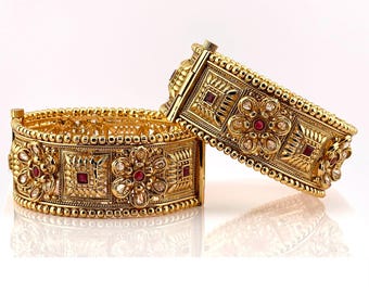 May include: Two gold-colored bangles with intricate floral and geometric designs. The bangles feature red and clear gemstone accents. One bangle is open, while the other is partially closed, showcasing the clasp. The jewelry is set against a white background.