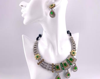May include: A silver and gold necklace with green gemstone accents. The necklace features a large, rectangular pendant with a floral design and a smaller, teardrop-shaped pendant. The necklace is displayed on a white mannequin.