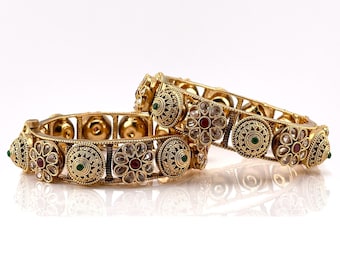 May include: Two gold bangles with intricate floral designs and red, green, and white gemstones.
