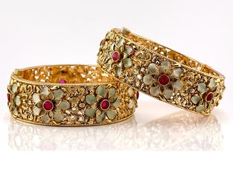 May include: Two gold bangles with intricate floral designs and red gemstones. The bangles are decorated with white enamel and have a delicate, ornate look.