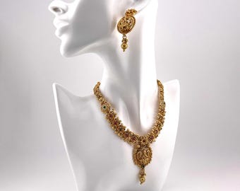 May include: A gold-toned necklace and earring set displayed on a white mannequin head. The necklace features a central pendant and is adorned with red, green, and gold accents. The earrings have a matching design. The jewelry is likely vintage or inspired by traditional designs.