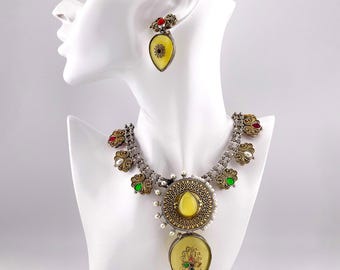 May include: A silver and gold necklace with a large yellow teardrop pendant. The pendant is surrounded by smaller stones in red, green, and white. The necklace is accented with pearl-like beads.