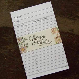 May include: A white library card notepad with lines for author, title, and borrower's name. A decorative band with the words "Library Card notepad" and floral accents is across the top. The notepad is on a dark wooden surface.