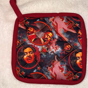 May include: A square quilted potholder with a dark background featuring a repeating pattern of vampire-themed portraits, red stars, and silhouettes. The fabric is edged in solid dark red trim and includes a matching fabric loop for hanging.