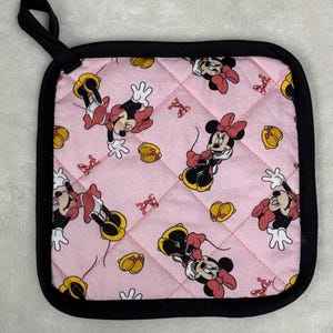 Könnte beinhalten: Rosa gesteppter Topflappen mit einem sich wiederholenden Muster von Minnie Mouse, Schleifen und gelben Knöpfen. Er hat einen schwarzen Rand und eine schwarze Schlaufe zum Aufhängen. Küchenzubehör.