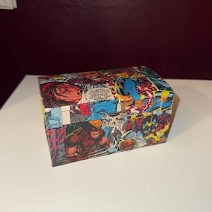 Peut inclure: Une boîte rectangulaire ornée de panneaux de bandes dessinées aux couleurs vives et aux scènes d'action. La boîte est posée sur une surface blanche, mettant en valeur l'œuvre colorée.