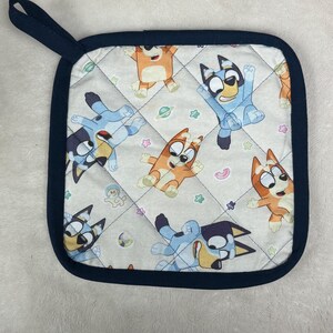 May include: Square quilted pot holder with a navy blue trim and hanging loop. The pot holder features a repeating pattern of cartoon dogs in various poses on a cream background. The dogs are blue and orange.