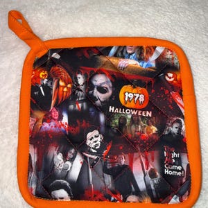 May include: A square quilted pot holder with an orange fabric border and a hanging loop. The surface features a collage of scenes from the 1978 Halloween film, including a pumpkin with the text 1978 HALLOWEEN and the phrase The Night He Came Home!