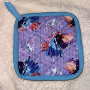 May include: A square quilted pot holder with a light blue fabric border and a matching hanging loop. The fabric features a repeating pattern of characters from the movie Frozen, including Elsa, Anna, and Olaf, set against a light purple background.