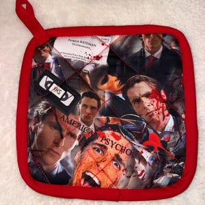 Könnte beinhalten: Ein quadratischer, gesteppter Topflappen mit rotem Stoffrand und Aufhängeschlaufe. Die Oberfläche zeigt eine Collage aus Szenen des Films American Psycho, darunter eine Visitenkarte von Patrick Bateman, ein VHS-Logo und blutige Spritzer.