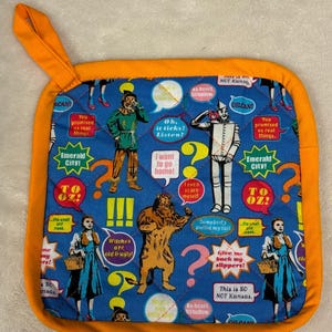 May include: A square, quilted pot holder with an orange border and loop. The blue center features characters and quotes from The Wizard of Oz, including the Tin Man, Scarecrow, and Dorothy. Text includes "Emerald City!" and "This is SO NOT Kansas."
