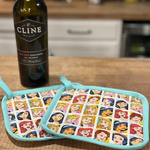May include: Two turquoise blue hot pads with a white background featuring a repeating pattern of Disney princess faces. The hot pads are on a wooden surface next to a bottle of Cline Old Vine Zinfandel wine.