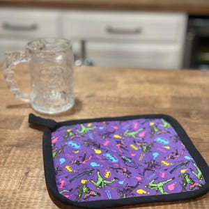 Puede incluir: Posavasos morado con borde negro. El posavasos tiene un patrón repetido de personajes de Batman en varias poses. Los personajes están en tonos de verde, rosa y amarillo.