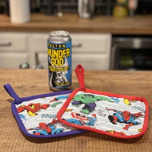 Puede incluir: Dos agarraderas de olla acolchadas de color rojo y morado con un tema de superhéroes. Las agarraderas de olla presentan imágenes de Spider-Man, Iron Man, Capitán América y Hulk. Las agarraderas de olla están sobre una superficie de madera junto a una lata de cerveza.