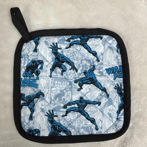 May include: Square quilted pot holder with a black border and hanging loop. The fabric features a comic book style print with blue Black Panther figures on a light blue and white background. The words "BLACK PANTHER" are visible.