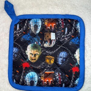 Può includere: Una presina quadrata trapuntata con bordo in tessuto blu e un occhiello per appenderla. Il tessuto presenta un motivo ripetitivo di Pinhead di Hellraiser, scatole puzzle dorate, catene argentate e schizzi di sangue rosso su sfondo nero.
