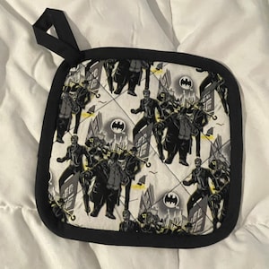 May include: Black and white pot holder with a Batman theme. The pot holder features a repeating pattern of Batman villains, including the Penguin, the Joker, and Catwoman. The pot holder has a black border and a black loop handle.