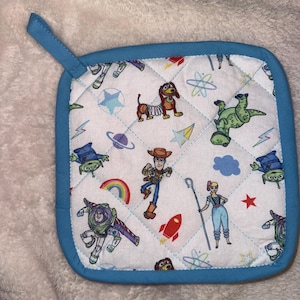 May include: A square quilted pot holder with a blue fabric border and a hanging loop. The white fabric features a repeating pattern of Toy Story characters including Woody, Buzz Lightyear, Bo Peep, Slinky Dog, Rex, and green aliens, alongside colorful stars, planets, and rockets.