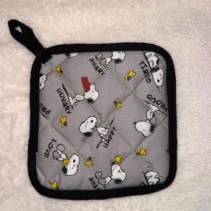 Pode incluir: Um pegador de panela quadrado cinza acolchoado com um padrão repetitivo de Snoopy e Woodstock. O tecido mostra Snoopy em várias poses com etiquetas de texto incluindo "ANGRY", "TIRED", "PROUD", "HAPPY", "HUNGRY" e "LOVE". Possui uma borda preta e uma alça.