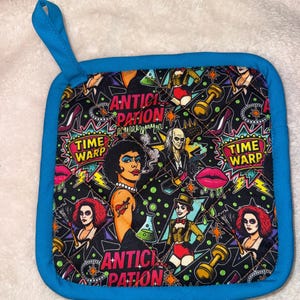 Puede incluir: Un agarrador de cocina cuadrado y acolchado con un borde de tela azul brillante y un lazo para colgar a juego. La tela presenta un fondo negro con un patrón colorido de personajes, labios rojos, mancuernas, tacones altos y el texto TIME WARP y ANTICIPATION.