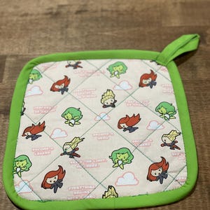 May include: Green and pink pot holder with a quilted design. It features a repeating pattern of cartoon superheroes with the text "Become a Hero", "Prepare to be Amazed", "Everything Begins with a Hero", "Second a Legend", "What's the Limit, Go There", and "Set a Good Example".