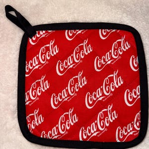 May include: A square quilted pot holder featuring a repeating white Coca-Cola logo pattern on a bright red background. The fabric is finished with a black fabric border and includes a small black loop at the top corner for convenient hanging storage.