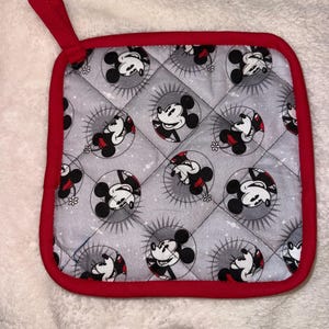 Puede incluir: Un agarrador de cocina cuadrado y acolchado con un patrón repetitivo de cabezas de Mickey Mouse dentro de marcos circulares sobre un fondo gris claro. El agarrador tiene un borde de tela rojo brillante e incluye una presilla para colgar a juego.