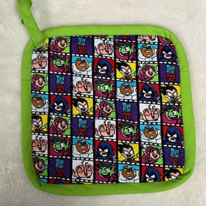 May include: Square, quilted pot holder with a lime green border and hanging loop. The center features a colorful pattern of Teen Titans Go! cartoon characters in a grid, with the words "TEEN TITANS GO!" in purple.
