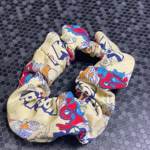 K&ouml;nnte beinhalten: Ein gelbes Haargummi mit einem bunten Cartoon-Print mit roten, blauen und orangenen Figuren. Der Stoff ist gerafft und elastisch, wodurch eine strukturierte, kreisf&ouml;rmige Form entsteht. Dieses Haaraccessoire ist f&uuml;r verschiedene Frisuren geeignet.