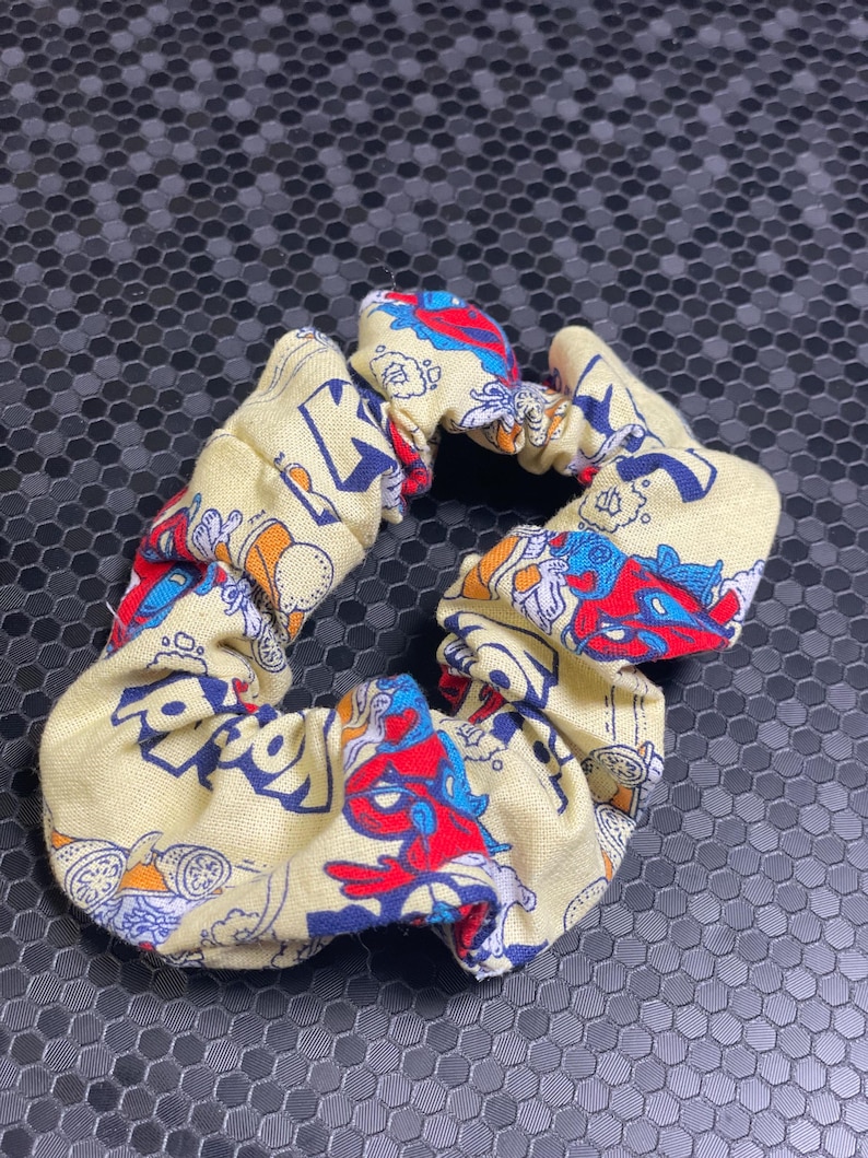 K&ouml;nnte beinhalten: Ein gelbes Haargummi mit einem bunten Cartoon-Print mit roten, blauen und orangenen Figuren. Der Stoff ist gerafft und elastisch, wodurch eine strukturierte, kreisf&ouml;rmige Form entsteht. Dieses Haaraccessoire ist f&uuml;r verschiedene Frisuren geeignet.