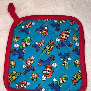 May include: A square quilted pot holder with a bright blue background featuring a repeating pattern of Mario and Luigi characters, fire flowers, and the Super Mario logo. The item has a red fabric border and a small red hanging loop at the top.