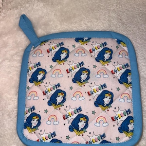 May include: A square quilted pot holder with a light pink background featuring a repeating pattern of a female superhero with blue hair, small rainbows, and the text KA-LOOM! in comic book style. The item has a solid light blue fabric border and loop.