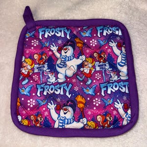Peut inclure: Un dessous de plat matelassé carré avec une bordure violette et une boucle de suspension. Le tissu présente un motif répétitif de Frosty le bonhomme de neige, un écureuil, un enfant et des oiseaux bleus sur un fond rose et violet avec le texte FROSTY.