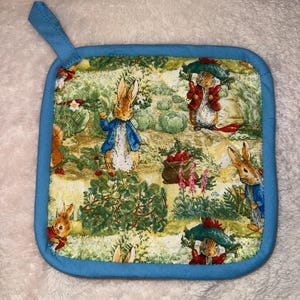 Puede incluir: Salvamanteles cuadrado con borde azul y lazo para colgar. La tela presenta un estampado caprichoso de conejos y otros personajes en un entorno de jardín. El salvamanteles está diseñado para proteger las superficies de los utensilios de cocina calientes.