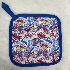 May include: Square blue pot holder with a Superman-themed fabric design. The fabric features Superman imagery and text including "Superman Saved Day" and "All American Hero". The pot holder has a blue border and a hanging loop.