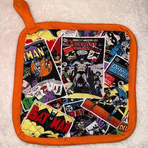 May include: A square quilted pot holder with a bright orange fabric border and a hanging loop. The centre features a colourful collage of vintage Batman comic book covers, including Detective Comics, with various illustrations of Batman and the Joker.