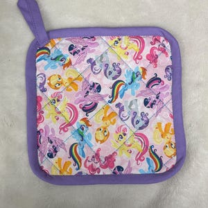May include: A square, quilted pot holder with a lavender border and hanging loop. The main fabric features a repeating pattern of colorful cartoon ponies on a pink background, with blue stitching creating a diamond pattern.