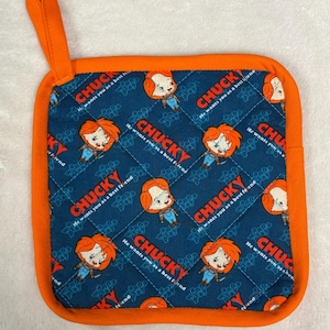 Puede incluir: Manopla de cocina cuadrada y acolchada con borde y lazo para colgar de color naranja. El centro presenta un patrón repetido del personaje de la muñeca Chucky y el texto "CHUCKY He wants you as a best friend" sobre un fondo azul oscuro.
