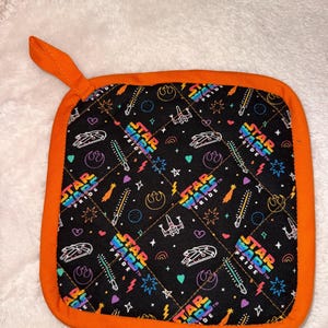 May include: A square quilted pot holder with a black background featuring a colourful Star Wars Pride pattern. The design includes rainbow-coloured Star Wars Pride logos, white line art of spaceships, lightsabers, and hearts, finished with an orange trim.