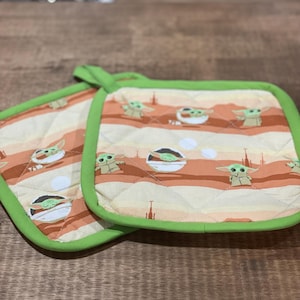 Può includere: Un paio di presine da forno verdi e arancioni con un design trapuntato. Le presine presentano un motivo ripetuto di un personaggio alieno verde del franchise di Star Wars.