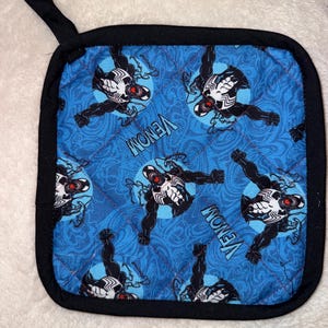 May include: A square quilted pot holder featuring a blue background with a swirling pattern and a repeating print of the character Venom. The design includes the text Venom in white lettering and a black fabric border with a small hanging loop.