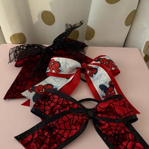 May include: Three hair bows with a Spider-Man theme. One bow features a red spiderweb design with black trim and black embellishments. Another bow has a red and white ribbon with Spider-Man graphics. The third bow has a black lace design.