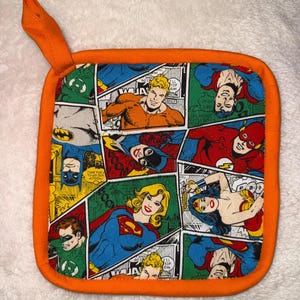 May include: A square pot holder with an orange fabric border and a hanging loop. The centre features a colourful comic book print displaying various DC Comics superheroes, including Batman, Superman, Wonder Woman, The Flash, and Green Lantern.