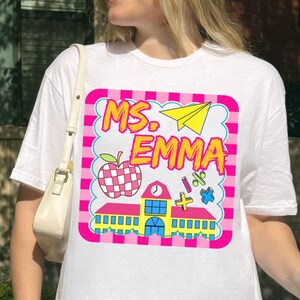May include: White t-shirt with a colorful graphic that says "Ms. Emma". The graphic includes a schoolhouse, apple, and other school-related images. The shirt is worn by a person.