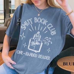 May include: Blue jean colored t-shirt with the text "Smut Book Club" in white, along with an illustration of a person on a book. The shirt has a relaxed fit and short sleeves. The text "One-Handed Reader" is also printed on the shirt.