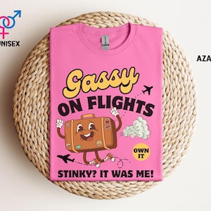 May include: Pink unisex t-shirt with the text "Gassy on Flights" and a cartoon suitcase character. The suitcase is brown with a smiling face and running legs. The shirt also has the text "Stinky? It was me!" and "Own it".