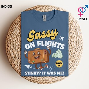 May include: Indigo blue unisex t-shirt with the text "Gassy on Flights" in yellow and white. A cartoon suitcase with arms and legs is featured in the design, along with airplane graphics and the phrase "Stinky? It was me!"