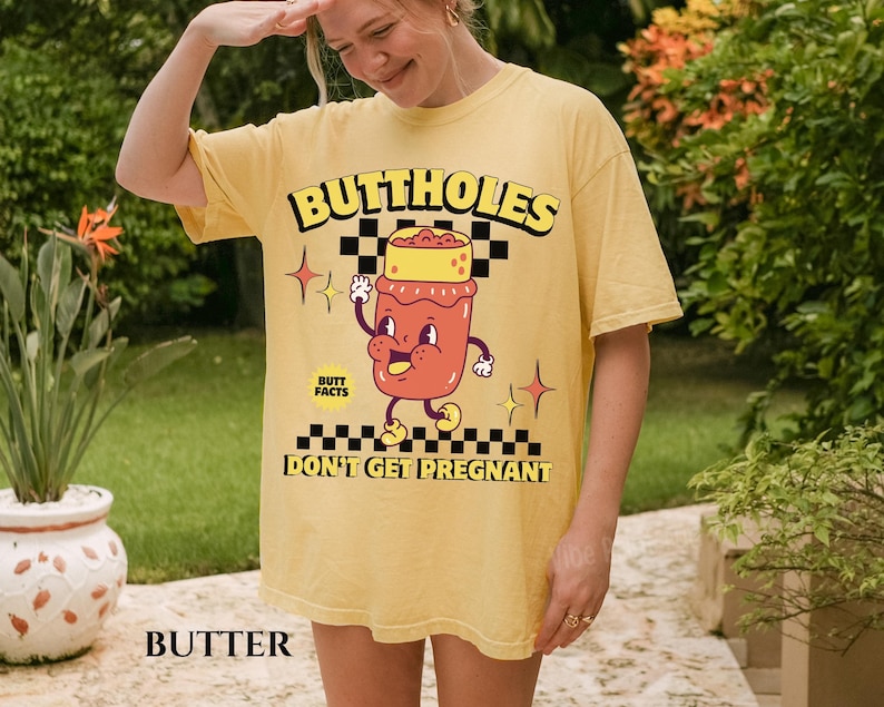 May include: Butter-colored t-shirt with the text "BUTTHOLES" in yellow and black. The shirt features a cartoon illustration of a jar with a face and the words "BUTT FACTS DON'T GET PREGNANT".