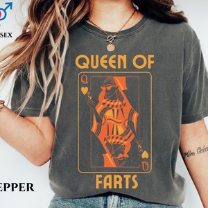 May include: Dark gray unisex t-shirt with a playing card design. The card features an orange queen wearing sunglasses, with the words "QUEEN OF FARTS" in a yellow font. The word "UNISEX" is above the shirt.