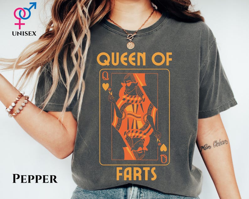 May include: Dark gray unisex t-shirt with a playing card design. The card features an orange queen wearing sunglasses, with the words "QUEEN OF FARTS" in a yellow font. The word "UNISEX" is above the shirt.